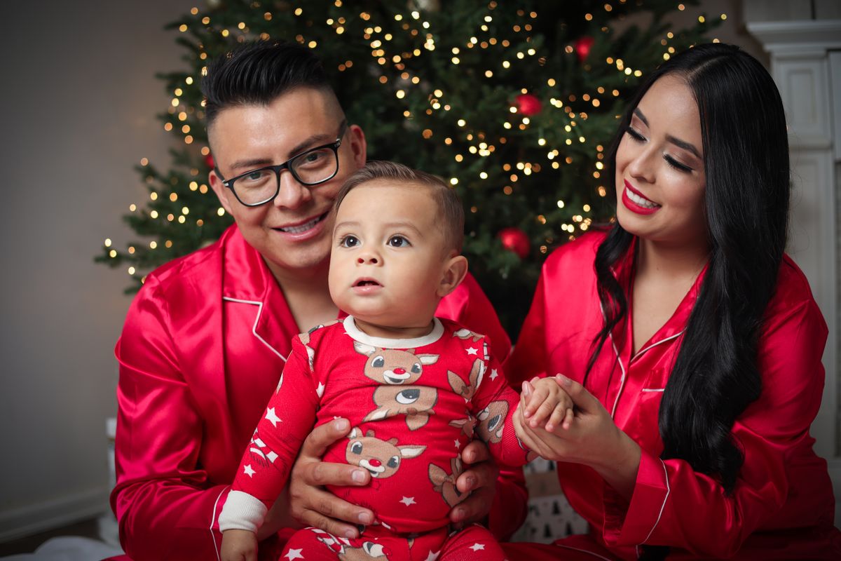 Casal com o bebê no colo, todos de pijama vermelho, diante de uma árvore de Natal iluminada